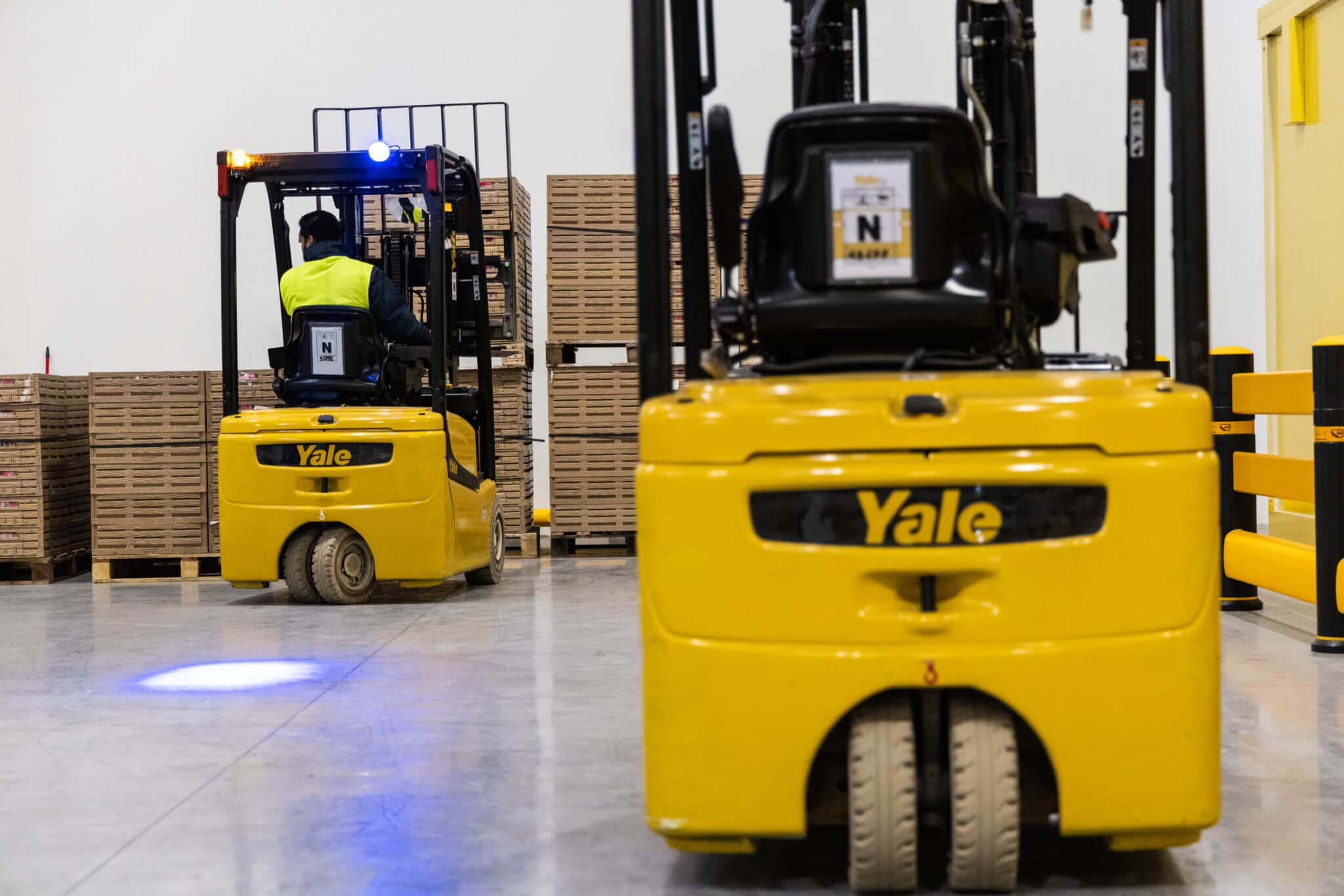 Two Yale forklifts, one parked and one reversing with blue safety light in use in warehouse.